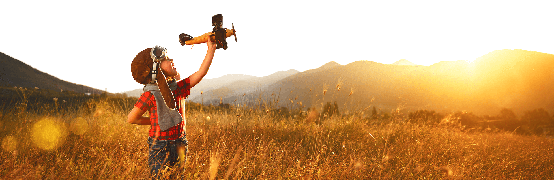 A young boy in an aviator cap and a red shirt holds a model airplane in a field under a bright sunrise