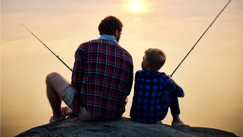 Father and young son sitting on a rock holding fishing poles with a sunset in the background