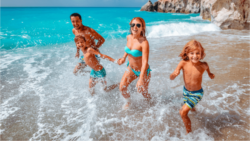 Mother and father run along a Caribbean beach with their two young boys. All wear blue swimsuits.