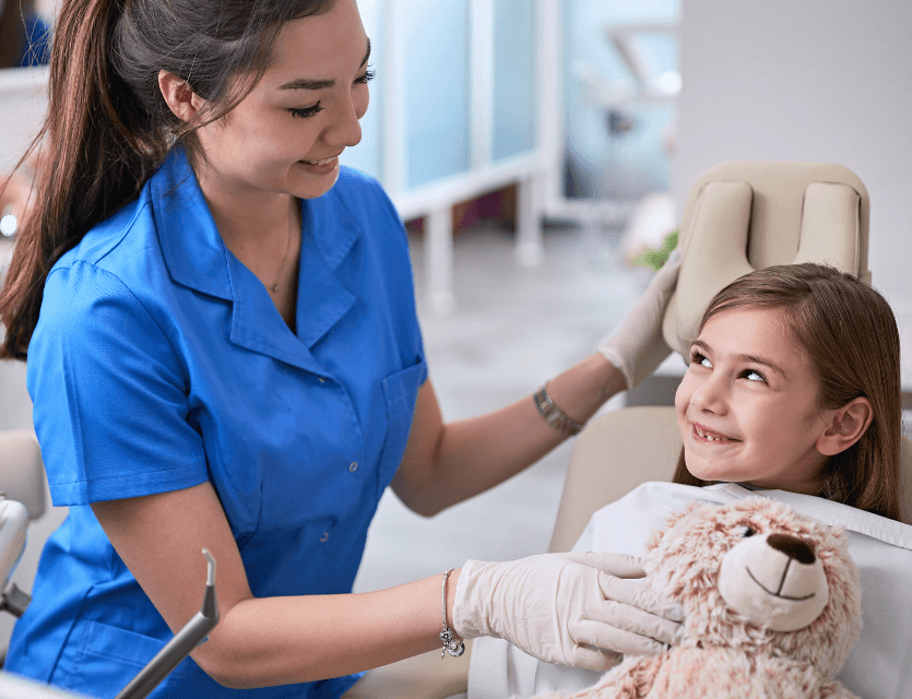 Young female dentist in blue scrubs smiling at a little girl in a dentists chair