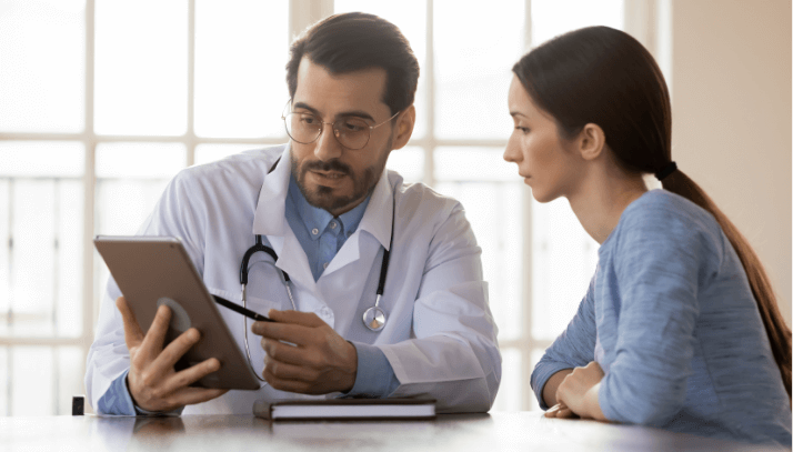 Male doctor in lab coat shares information on a tablet with a young female patient in blue shirt