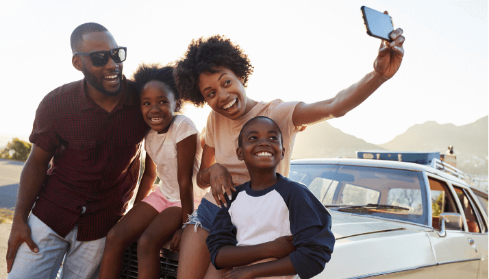 Dad, mom, and two young kids on a road trip taking a selfie picture on the hood of their car