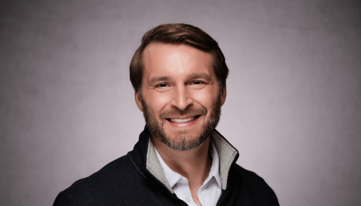Male business professional in a white shirt under a dark blue sweater against a light brown background